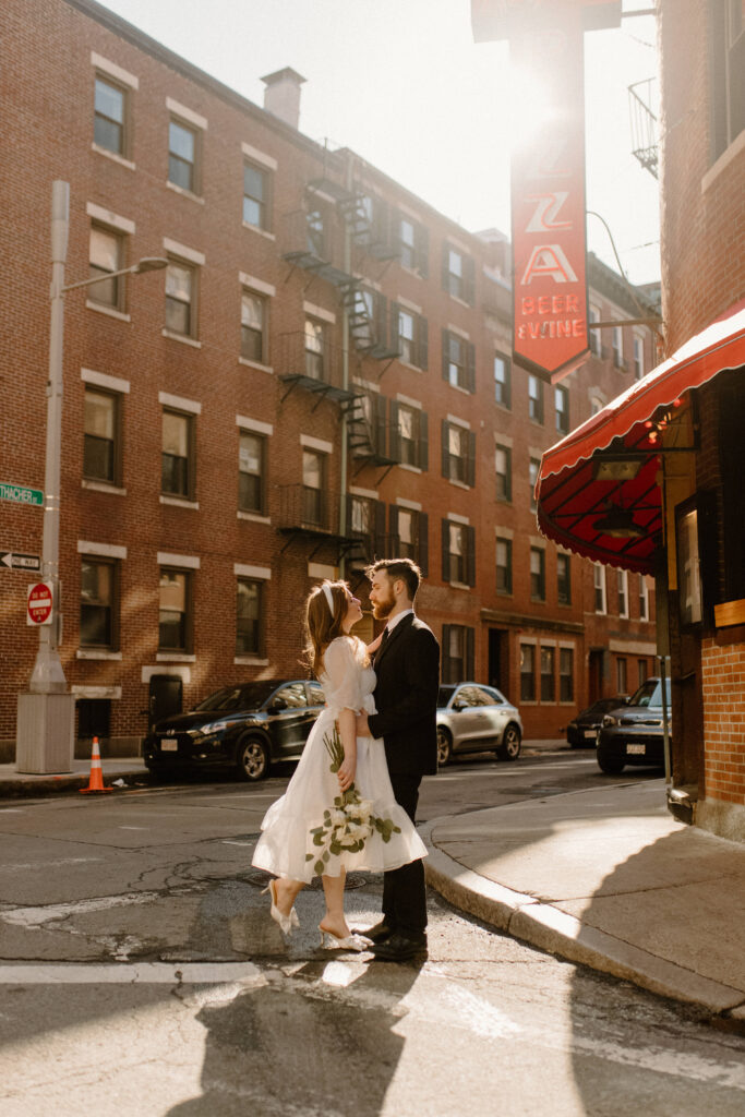A bride and groom during their elopement holding flowers outside a pizza shop in the city after learning wedding planning tips
