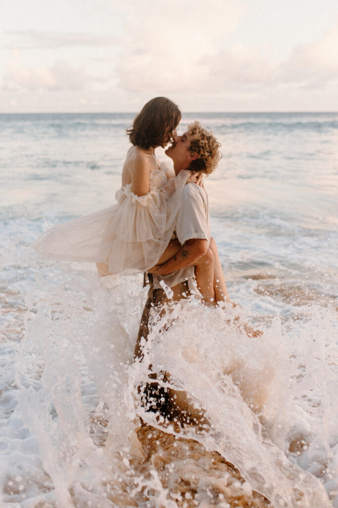 A newly engaged couple enjoying their engagement session on a Hawaii beach all snuggled up in a wave after learning wedding planning tips 