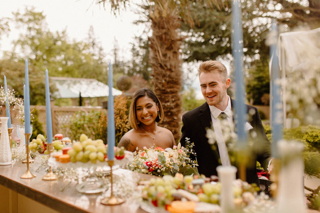 A bride and groom smiling at their wedding catering table with flowers and fruits and desserts after learning wedding planning tips 