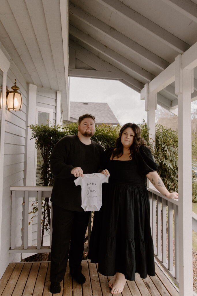 Couple on their porch holding a baby onsie for their in home pregnancy announcement photoshoot.