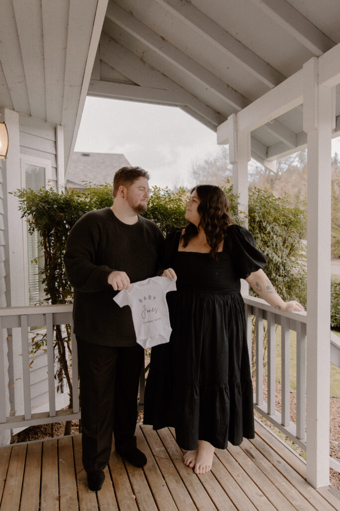 Couple on their porch holding a baby onsie for their in home pregnancy announcement photoshoot.