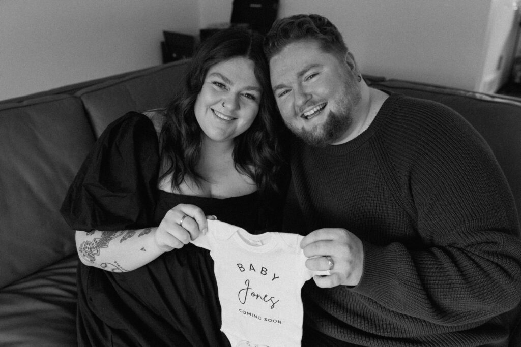 Couple smiling posing with a onsie to announce their pregnancy.