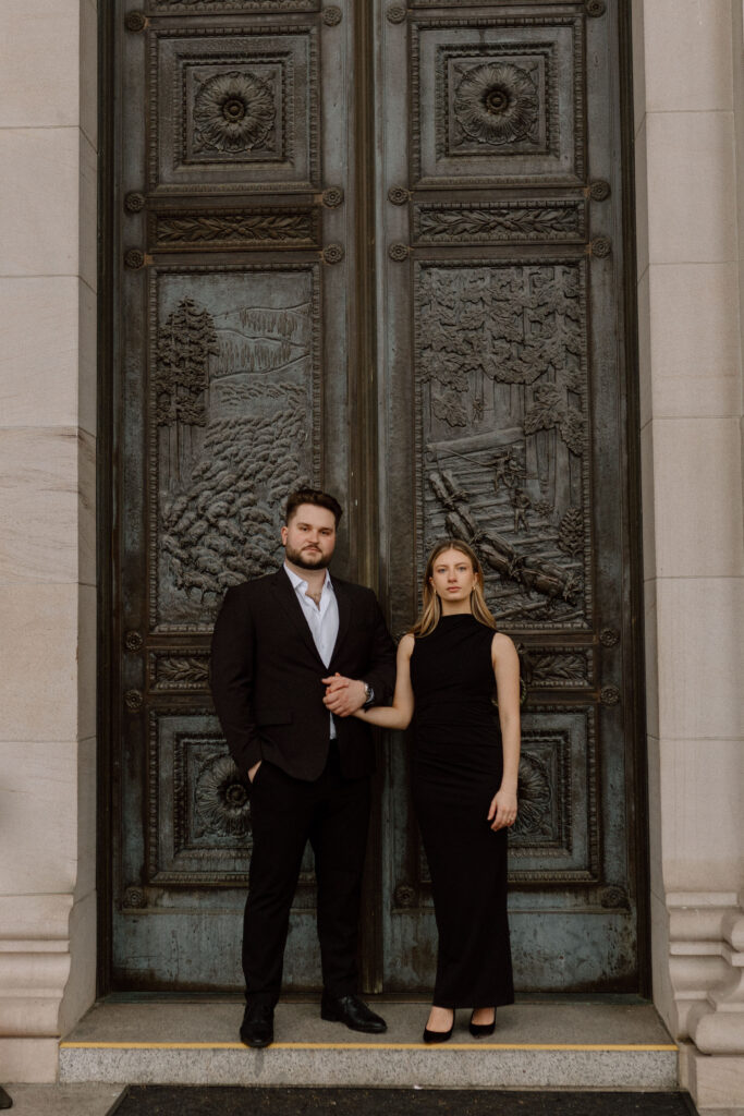 A couple dressed up standing in front of fancy doors durning their classic engagement photoshoot.