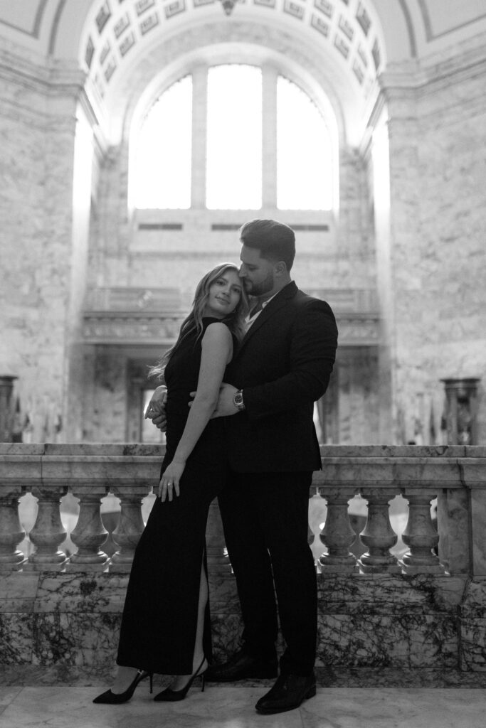 Engaged couple inside Washington Capitol building with arched windows behind them.