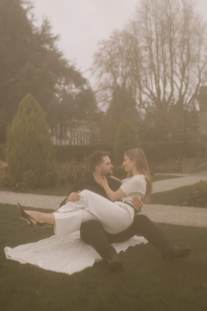 A couple sitting on a blanket in the middle of a garden with her on his lap.