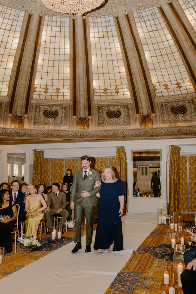Groom and mother walk down the wedding aisle 